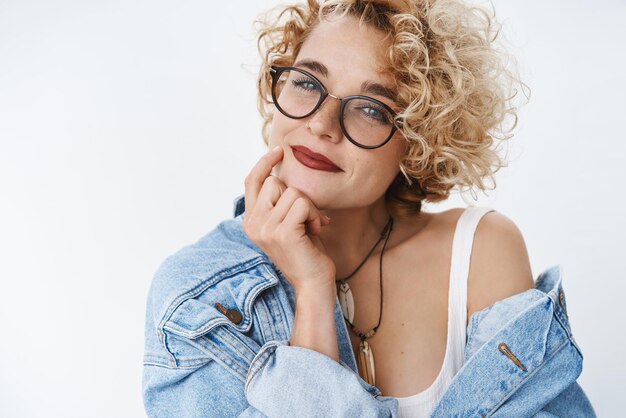 Closeup shot of romantic and selfassured modern girl with blond hairstyle and glasses smiling flirty and tender holding fingers on jawling gazing cute at camera over white background