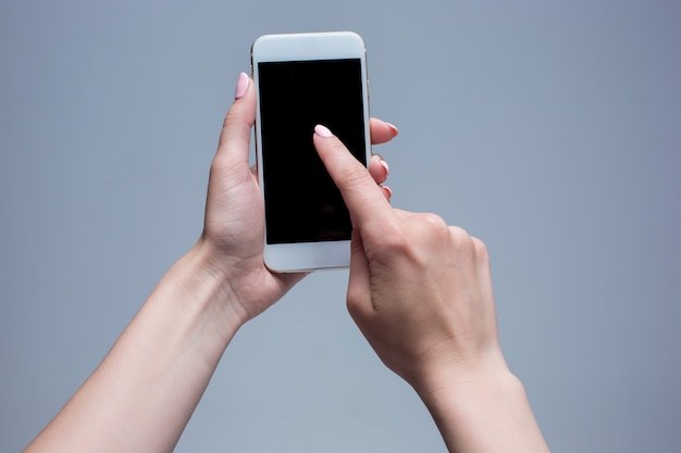 Closeup shot of a woman typing on mobile phone on gray background. Female hands holding a modern smartphone and pointing with figer. Blank screen to put it on your own webpage or message.