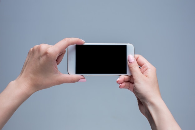 Closeup shot of a woman typing on mobile phone on gray background. Female hands holding a modern smartphone and pointing with figer. Blank screen to put it on your own webpage or message.