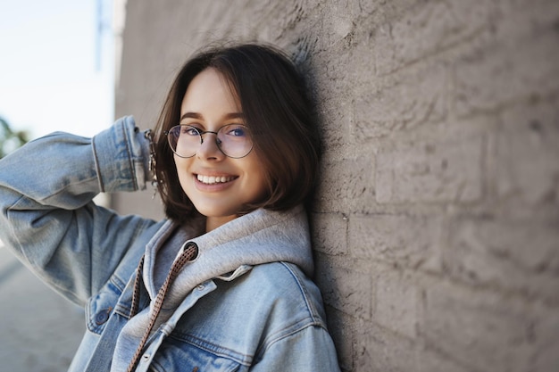 Closeup side portrait of adorable young queer woman in glasses lean brick wall outdoors on street looking and smiling camera happy feeling upbeat walking along city nice spring weather