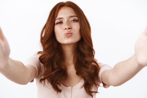 Closeup silly attractive glamour young tender redhead woman in striped tshirt with curly hairstyle extend arms and holding camera both hands using smartphone to video call pouting blowing kiss