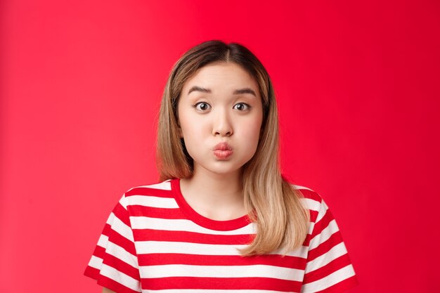 Closeup silly cute asian blond girl pouting making playful face fold lips hold breath stand red back...