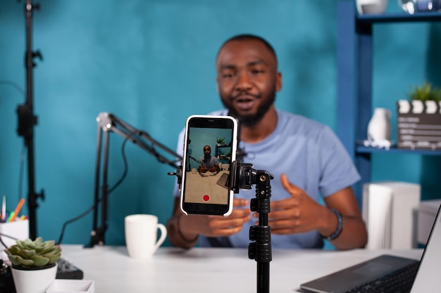 Closeup of smartphone filming influencer sitting down at desk with laptop smiling interacting with fans. Selective focus on live video podcast setup recording content creator moving hands gesturing.
