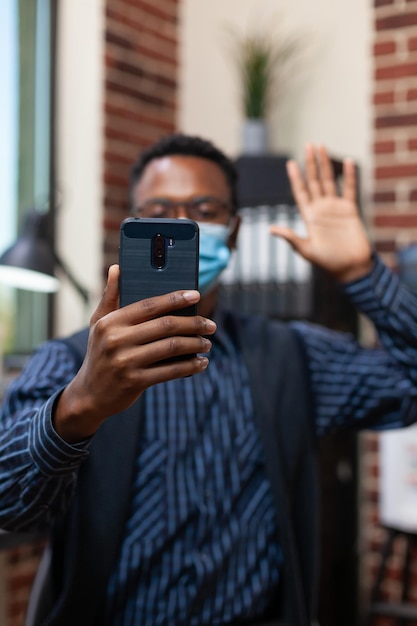 Closeup of smartphone of startup employee wearing face mask waving hello in online interview presenting himself. Detail view of mobile phone held by entrepreneur in video call conference.