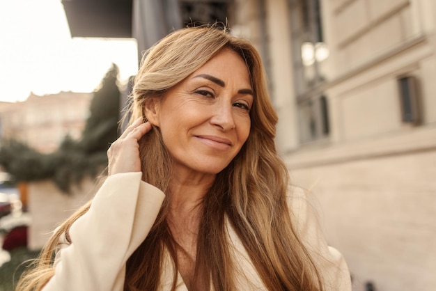 Closeup of smiling fairskinned adult woman looking at camera while arranging blond hair behind her ear People sincere emotions lifestyle concept