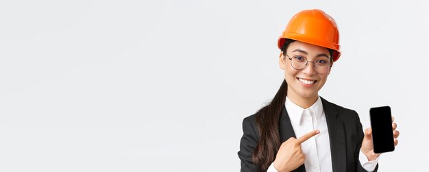 Closeup of smiling professional female asian construction engineer architect in suit and safety mask introduce application pointing finger at smartphone display white background