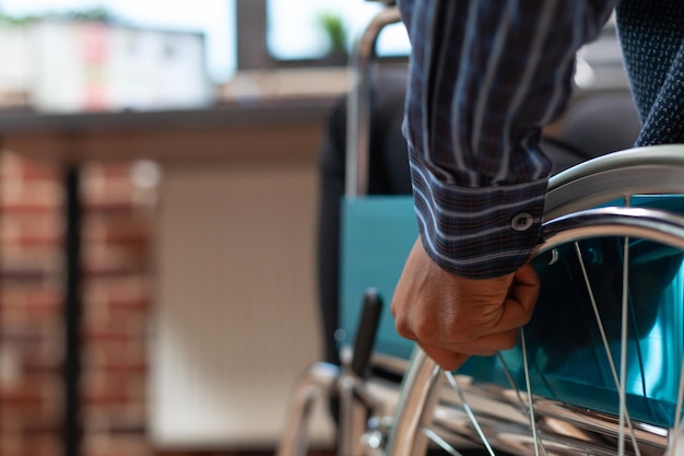 Closeup of startup employee hand holding rim of wheelchair wheel to move in front of desk with laptop with sales charts. Focus on african american man living with disability moving around office.