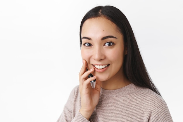 Closeup studio shot attractive asian girl with long dark hair wear sweater touching cheek and smiling delighted surprised with someone mentioned her skincare progress stand white background