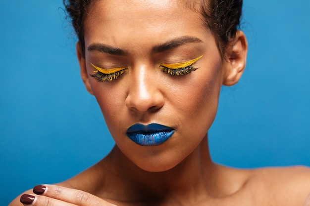 Closeup stylish relaxed mixed-race woman with colorful cosmetics on face posing on camera with closed eyes, isolated over blue wall