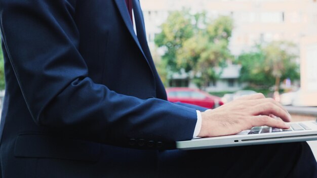 Closeup of successful leader hand typing marketing strategy on laptop computer keyboard sitting on bench in front of startup company office. Executive manager planning business presentation