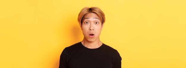 Closeup of surprised speechless asian guy drop jaw and raising eyebrows amazed standing over yellow