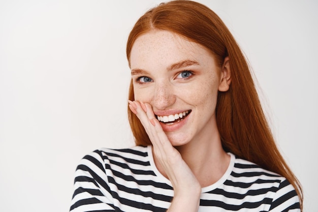 Primo piano di una ragazza adolescente dai capelli rossi con lentiggini e occhi azzurri, guardando davanti, toccando la pelle pulita e sana sul viso e sorridendo