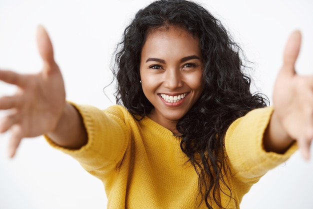 Closeup tender and cute pretty african american curlyhaired woman in yellow sweater stretchin hands forward smiling and gazing camera with love and care to give hug cuddle friend