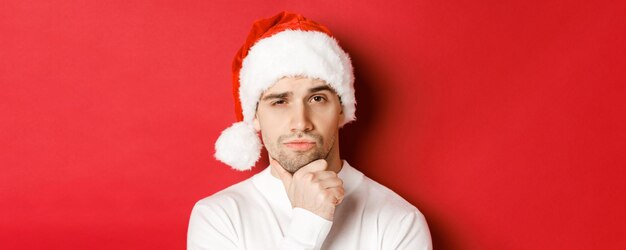 Closeup of thoughtful handsome man in santa hat frowning and looking at camera thinking about someth...