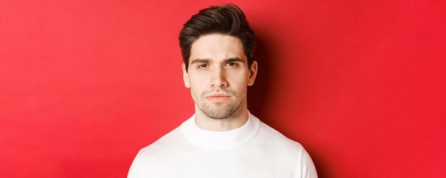 Closeup of thoughtful seriouslooking man in white sweater standing over red background