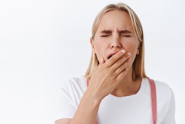 Closeup tired yawning cute blond woman in tshirt cover opened mouth with palm as falling asleep feeling fatigue or exhaust close eyes wake up early morning wanna coffee white background