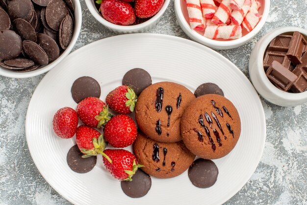 근접 촬영 상위 뷰 쿠키 딸기와 회색 흰색 테이블에 사탕 딸기와 초콜릿으로 둘러싸인 흰색 타원형 접시에 둥근 초콜릿