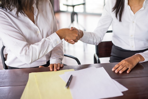 Closeup of two business women shaking hands and sitting at desk. 