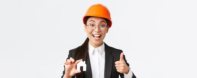 Closeup of upbeat professional female asian engineer architect in helmet and business suit encourage contact her company for renovation and design showing house and thumbsup