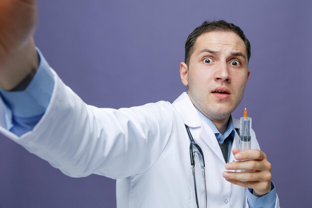 Крупный план испуганного молодого врача-мужчины в медицинском халате и стетоскопе на шее, смотрящего в камеру, показывающую шприц с иглой, протягивающей руку к камере, делающей селфи