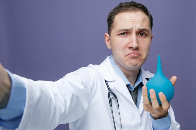 closeup view of unpleased young male doctor wearing medical robe and stethoscope around neck looking at camera showing enema stretching hand out towards camera taking selfie