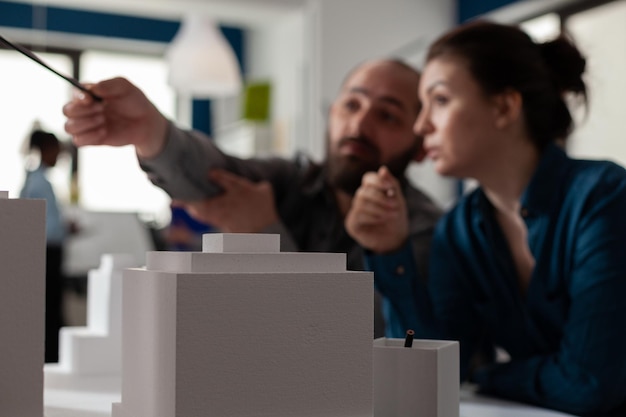 Closeup of white foam architectural scale model of buildings in front of architects team pointing at residential project. detail of urban development maquette on design table.