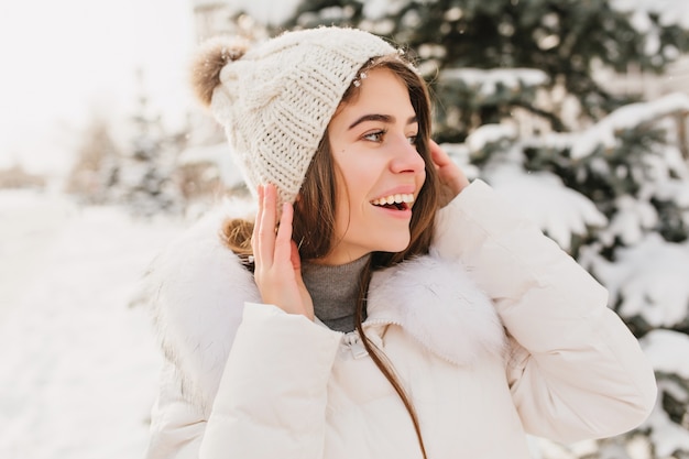 Крупным планом зимний портрет молодая красивая женщина в белой связанной шляпе, выражающей сторону на улице, полной снега.