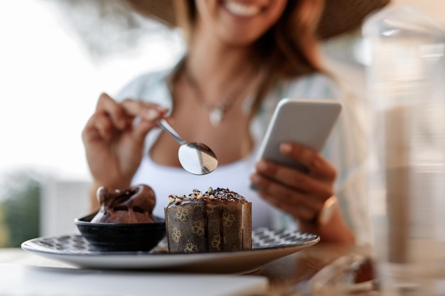 Крупный план женщины, использующей смартфон во время поедания торта в кафе. Фокус на переднем плане