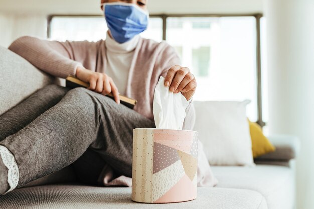 Closeup of woman wearing face mask and using paper tissue while sitting on the sofa at home