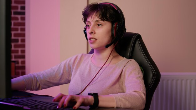 Closeup of woman with gaming headset playing multiplayer first person shooter streaming gameplay on professional pc setup. Gamer girl looking at computer screen while talking to team in tournament.