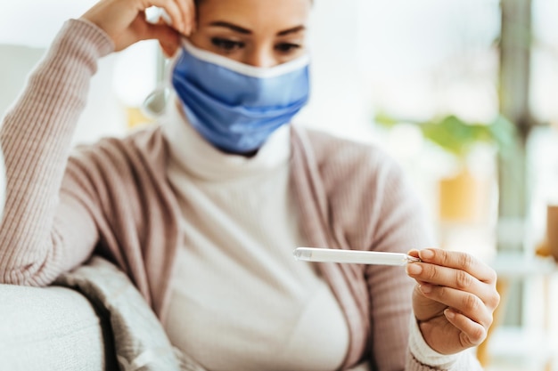 Primo piano della donna con maschera facciale protettiva che utilizza il termometro e misura la sua temperatura a casa