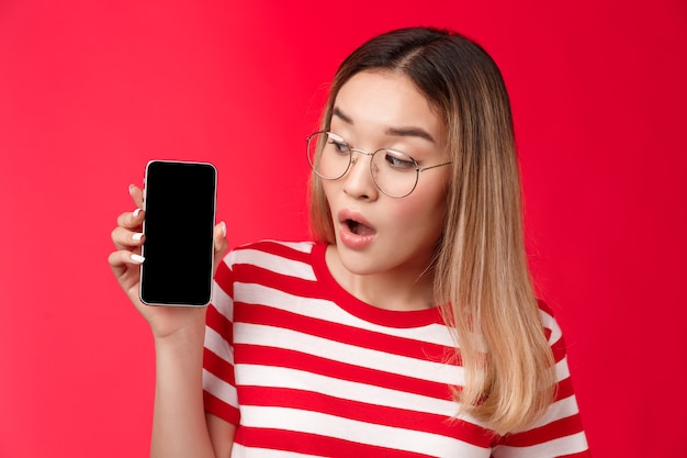 Closeup wondered surprised attractive asian blond girl wear glasses hold smartphone looking impresse...