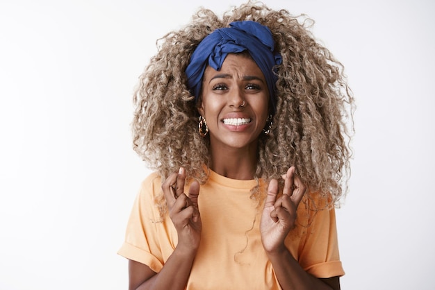 Closeup worried and anxious troubled young hopeless africanamerican curly blond woman in orange tshirt headband clench teeth desperate look camera cross fingers good luck praying