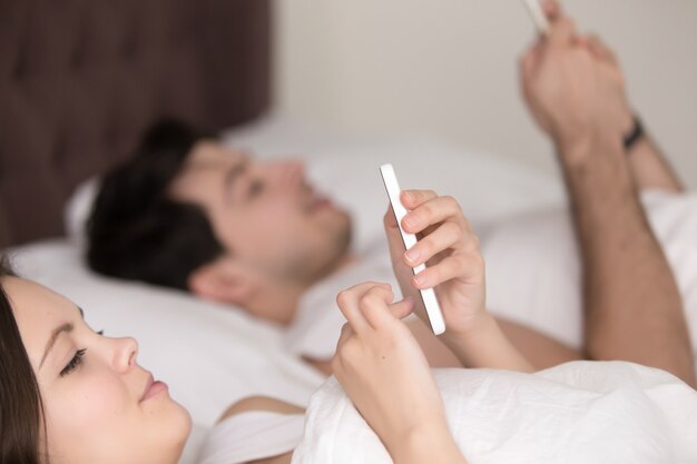 Closeup of young couple using mobile smartphones lying in bed