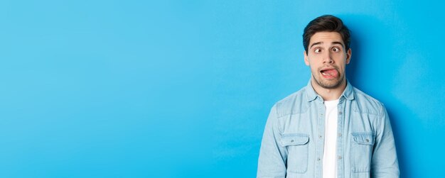 Closeup of young man making funny expressions showing tongue and looking at camera standing over blu