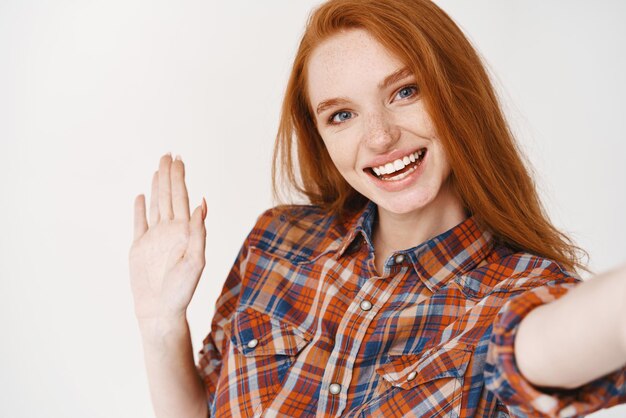 Closeup of young woman with red long hair and blue eyes taking selfie and saying hi Girl blogger waving hand and smiling at camera