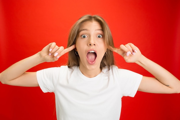 Closing ears. Portrait of young caucasian woman with bright emotions on red  wall with copyspace. Blonde model. 