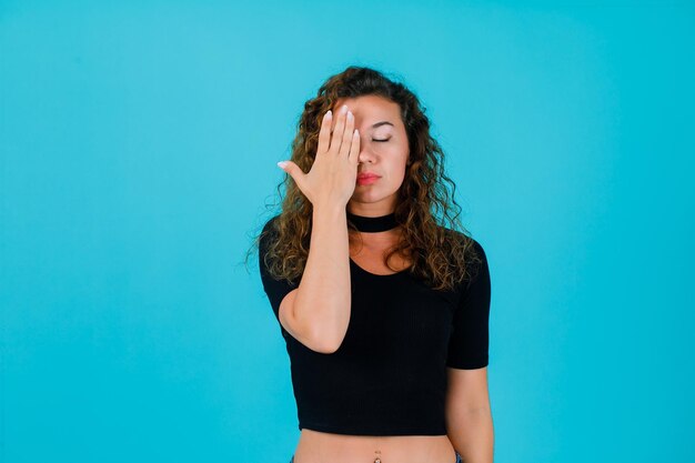 Clossed eyes girl is covering right side of face with hand on blue background
