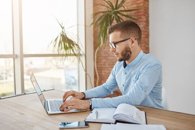 Closw up portrait of adult concentrated unshaven male company accountent in glasses and shirt sitting in comfortable office working on laptop computer.