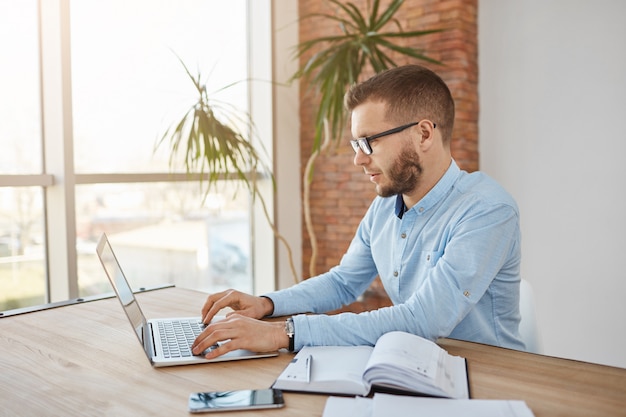 Closw up portrait of adult concentrated unshaven male company accountent in glasses and shirt sitting in comfortable office working on laptop computer.