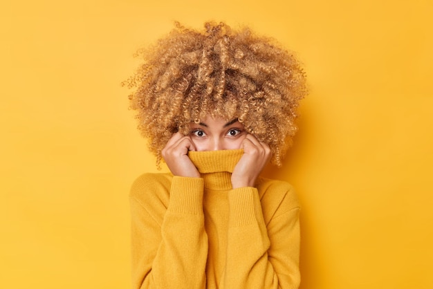 Abbigliamento per la stagione fredda la donna dai capelli piuttosto riccia copre la bocca e il naso con il colletto di un maglione lavorato a maglia alla moda che guarda sorprendentemente le pose della fotocamera su uno sfondo giallo che si riscalda durante l'inverno