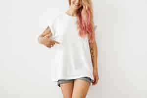 Foto gratuita abbigliamento, design, stile, moda e pubblicità. ritratto di irriconoscibile donna sottile hipster con riflessi di capelli rosa e braccia tatuate sorridente, puntando il dito a t-shirt vuota