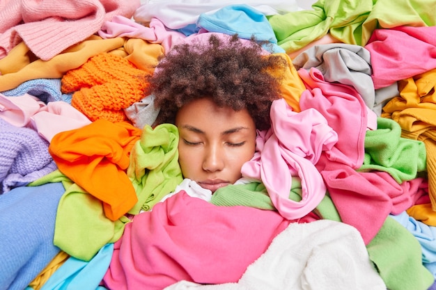 Clothing dilemma. Curly haired woman buried in stack of multicolored clothes