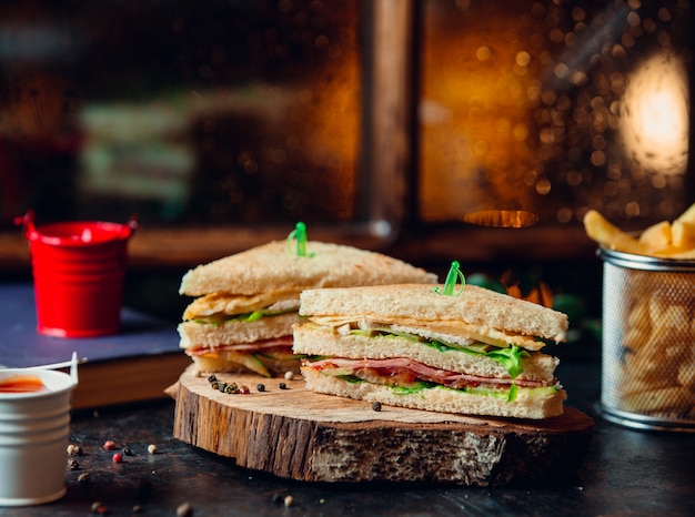 Club sandwich with ham, lettuce, tomato, cheese, and fries on wooden board