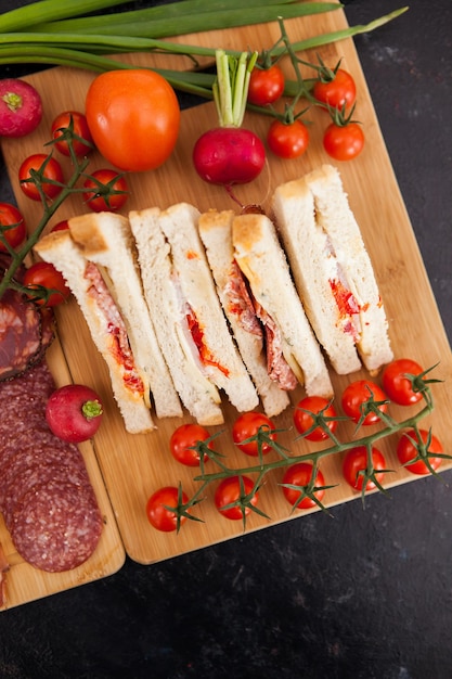 Club sandwiches lying on wooden board next to radish, cherry tomatoes and green onion