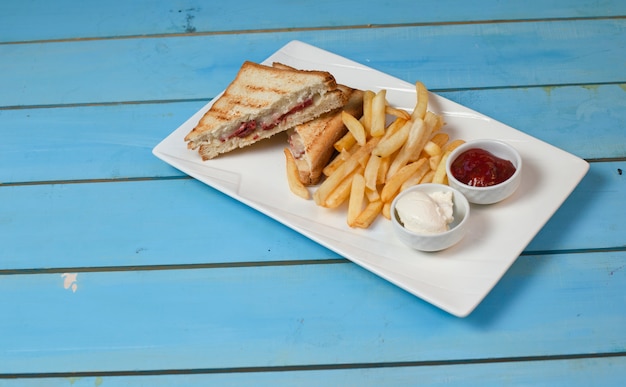 Club sandwiches served with fried potatoes in white plate with ketchup and mayonnaise on blue table