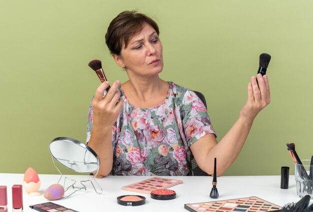 Clueless adult caucasian woman sitting at table with makeup tools holding and looking at makeup brushes isolated on olive green wall with copy space