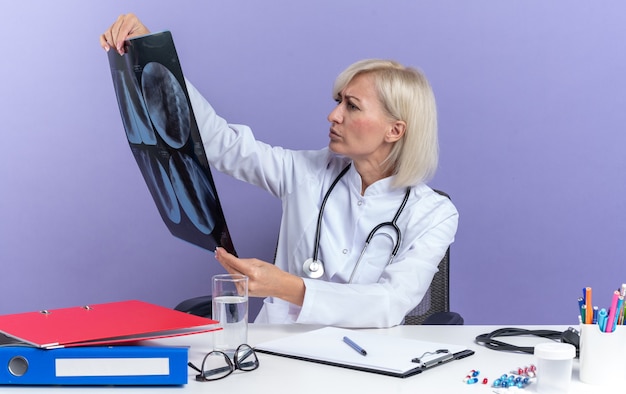 clueless adult female doctor in medical robe with stethoscope sitting at desk with office tools holding and looking at x-ray result isolated on purple wall with copy space