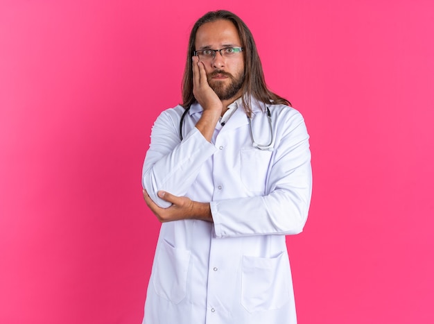 Clueless adult male doctor wearing medical robe and stethoscope with glasses keeping hand on face and on elbow looking at camera isolated on pink wall with copy space