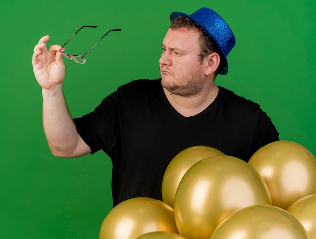Clueless adult slavic man holds and looks at optical glasses wearing blue party hat standing with helium balloons 